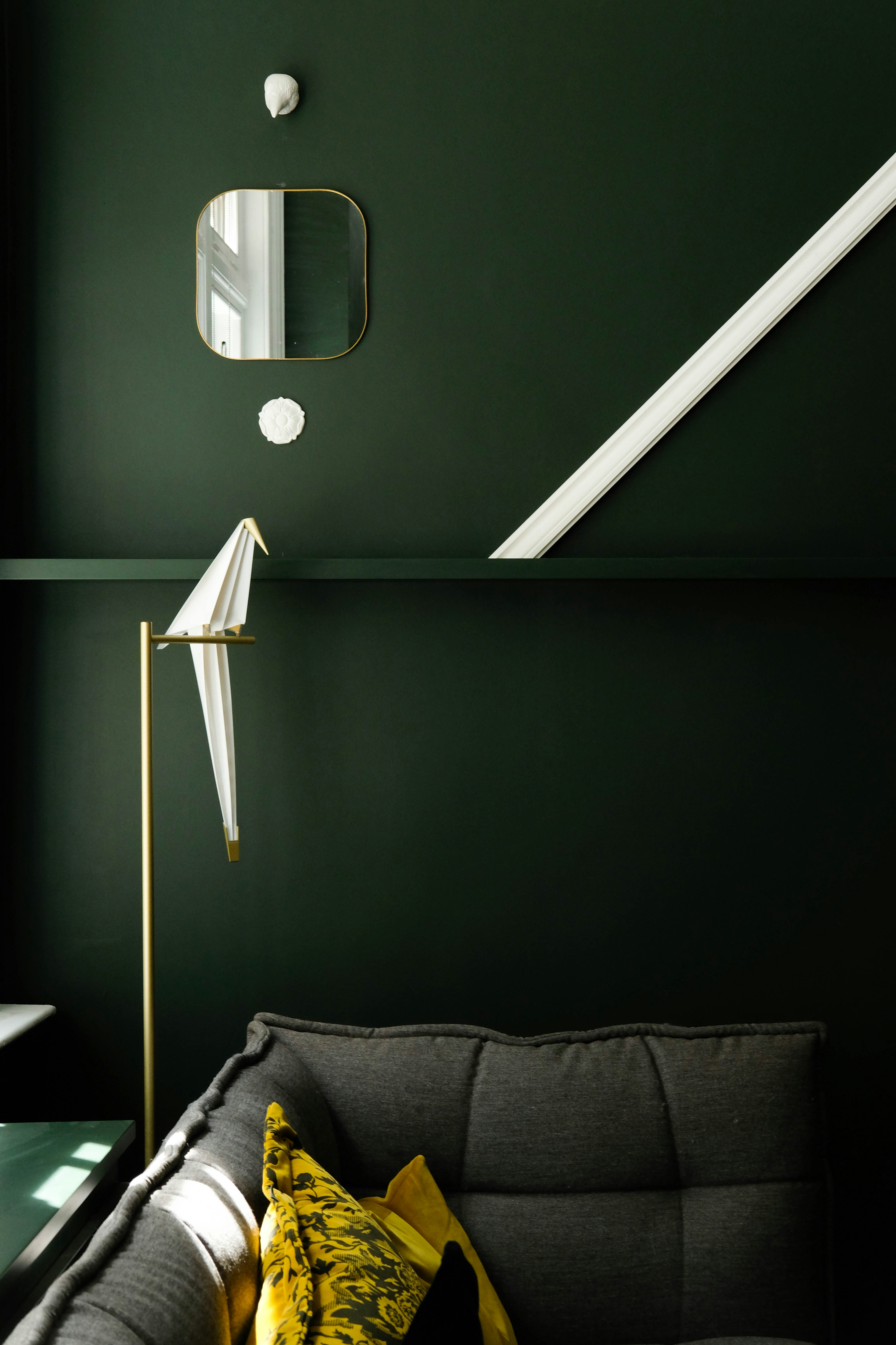 A modern interior with a dark green wall, gold-framed mirror, sculptural ornaments, and an origami-inspired bird lamp. A gray couch with yellow and black pillows adds contrast.